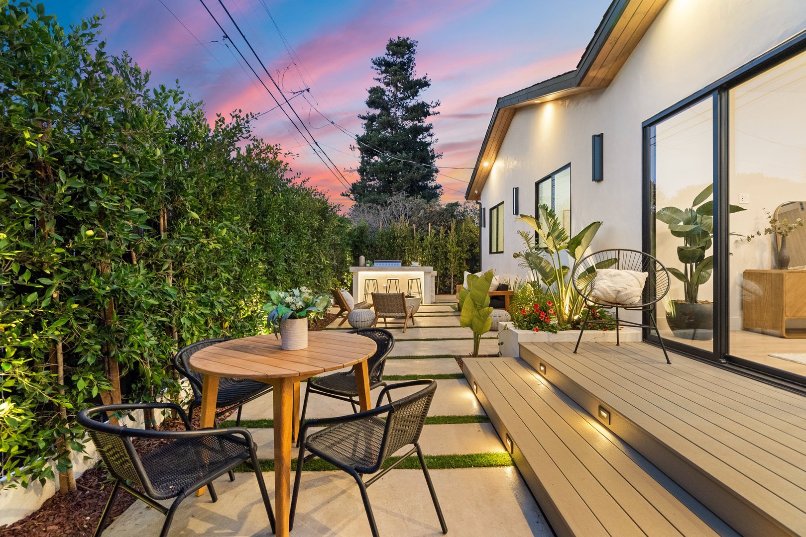 Modern backyard patio with stylish furniture and ambient lighting at sunset, surrounded by lush greenery.