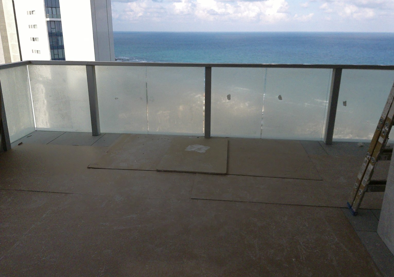 A balcony with a glass railing under construction, overlooking the ocean with a partially cloudy sky.