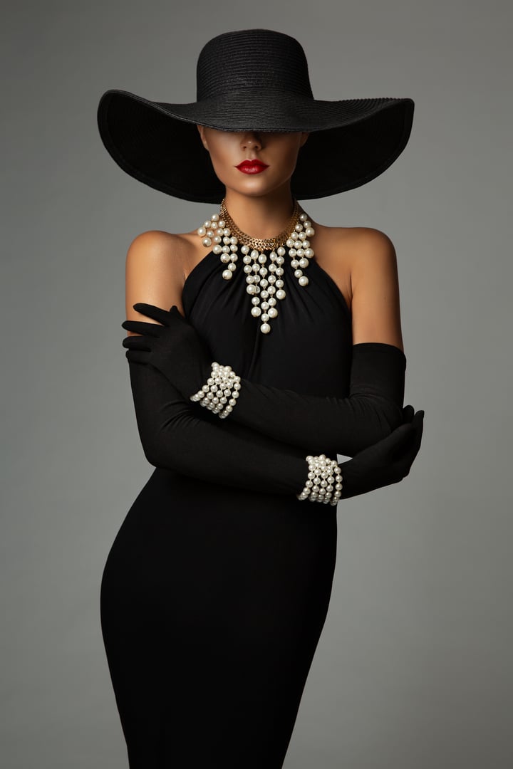 Elegant Lady in Black Dress wearing Pearl Jewellery Necklace and Wide Brim Hat. Fashion Woman in Cocktail Form Fitted Gown and Gloves with Bracelets over Gray. Stylish Model with hidden Face