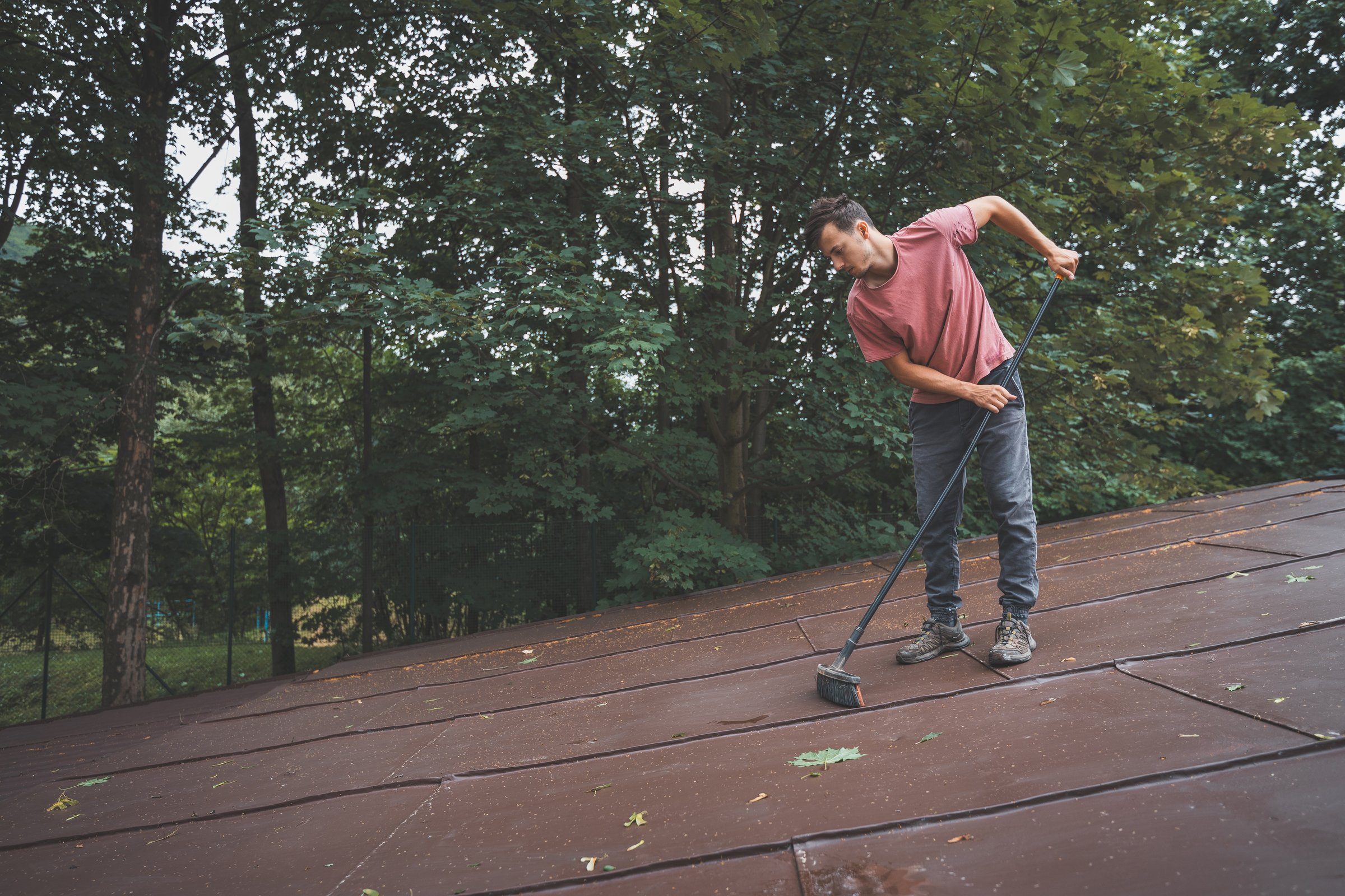 Man is cleaning a slanted roof with a long broom, surrounded by trees. The outdoor setting features lush greenery, with the individual focused on his task of removing debris from the roof.