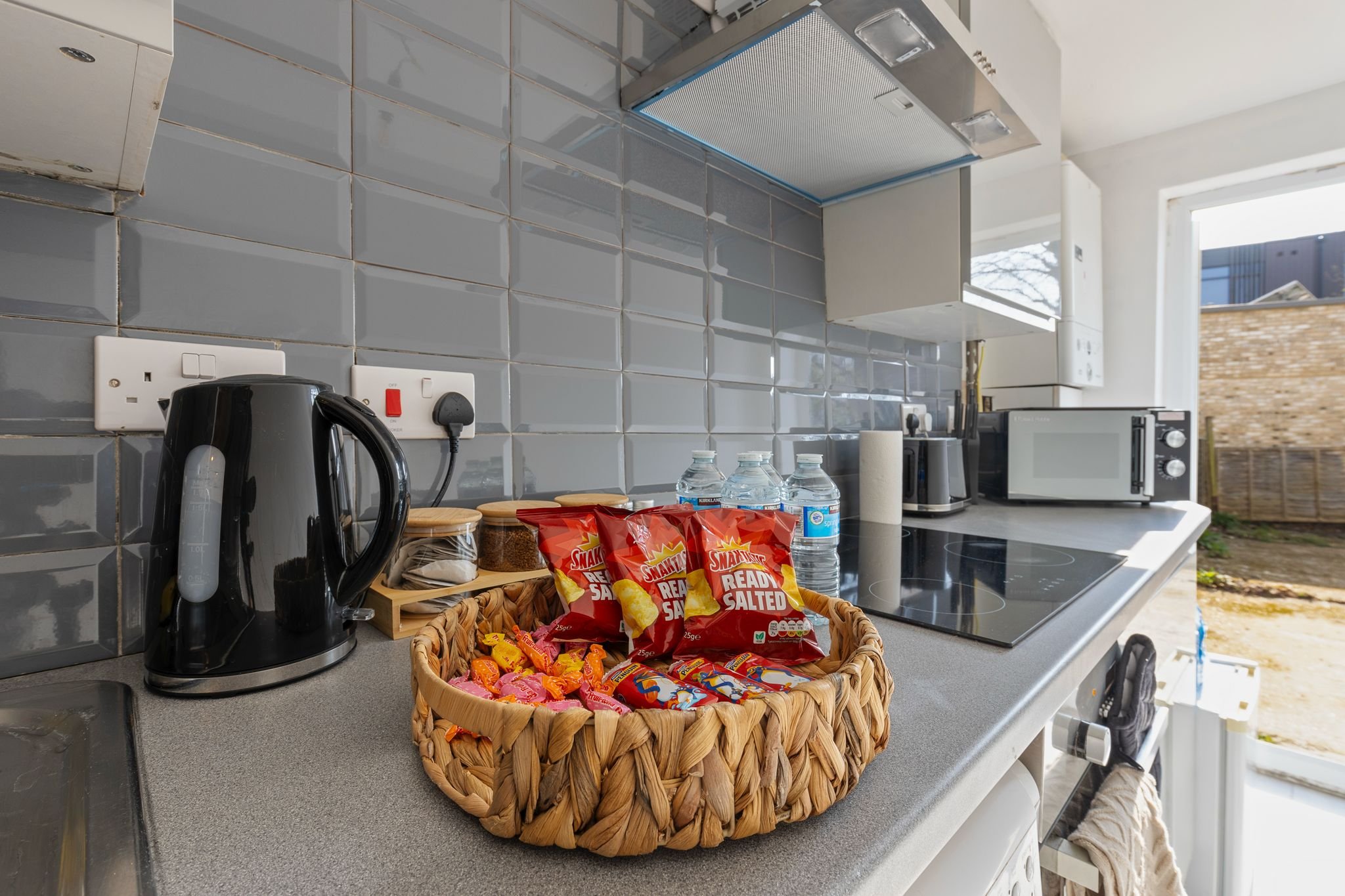 Kitchen counter with snack basket, kettle, two water bottles, and a microwave against a gray tiled backsplash.