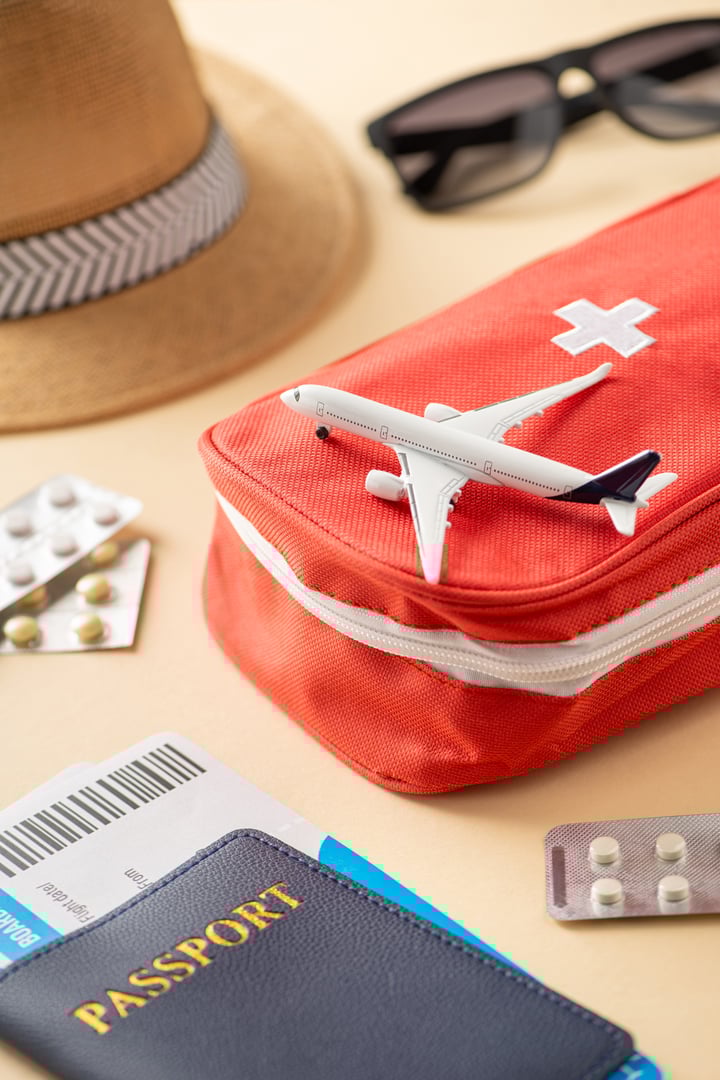 Travel-themed vertical arrangement featuring a first aid kit, passport, sunglasses, pills, and a toy airplane representing important travel essentials