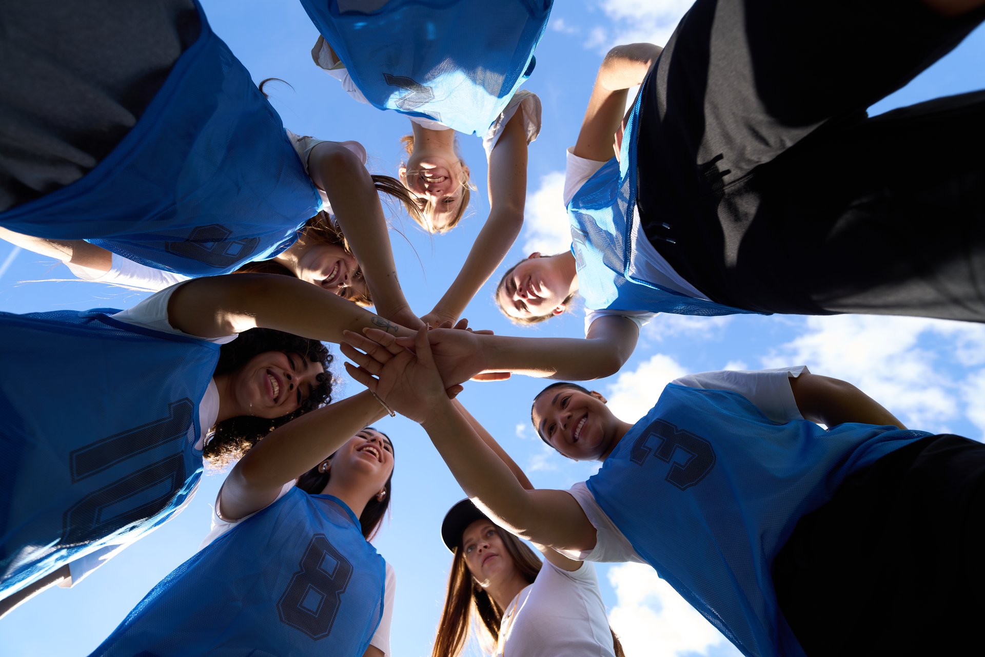 Diverse young women soccer team huddling, stacking hands in the center, showing unity and support on a bright day