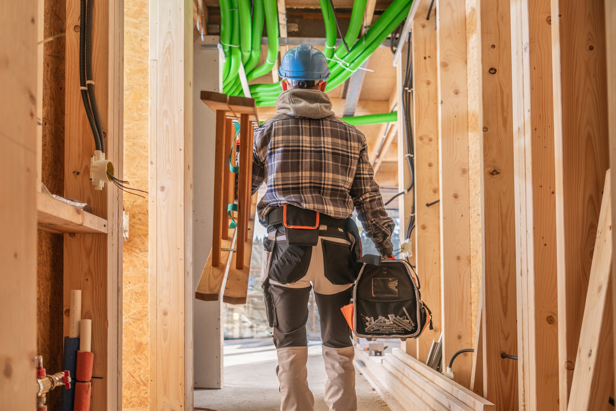 A construction worker carries tools while walking through a wooden framework with green pipes overhead, highlighting an active construction site.