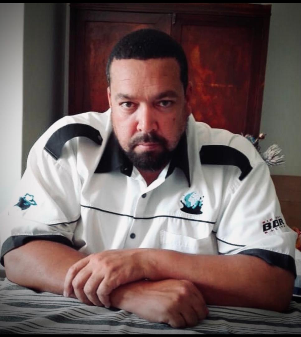 Man sitting at a table with hands clasped, wearing a white and black shirt, in front of a wooden cabinet.