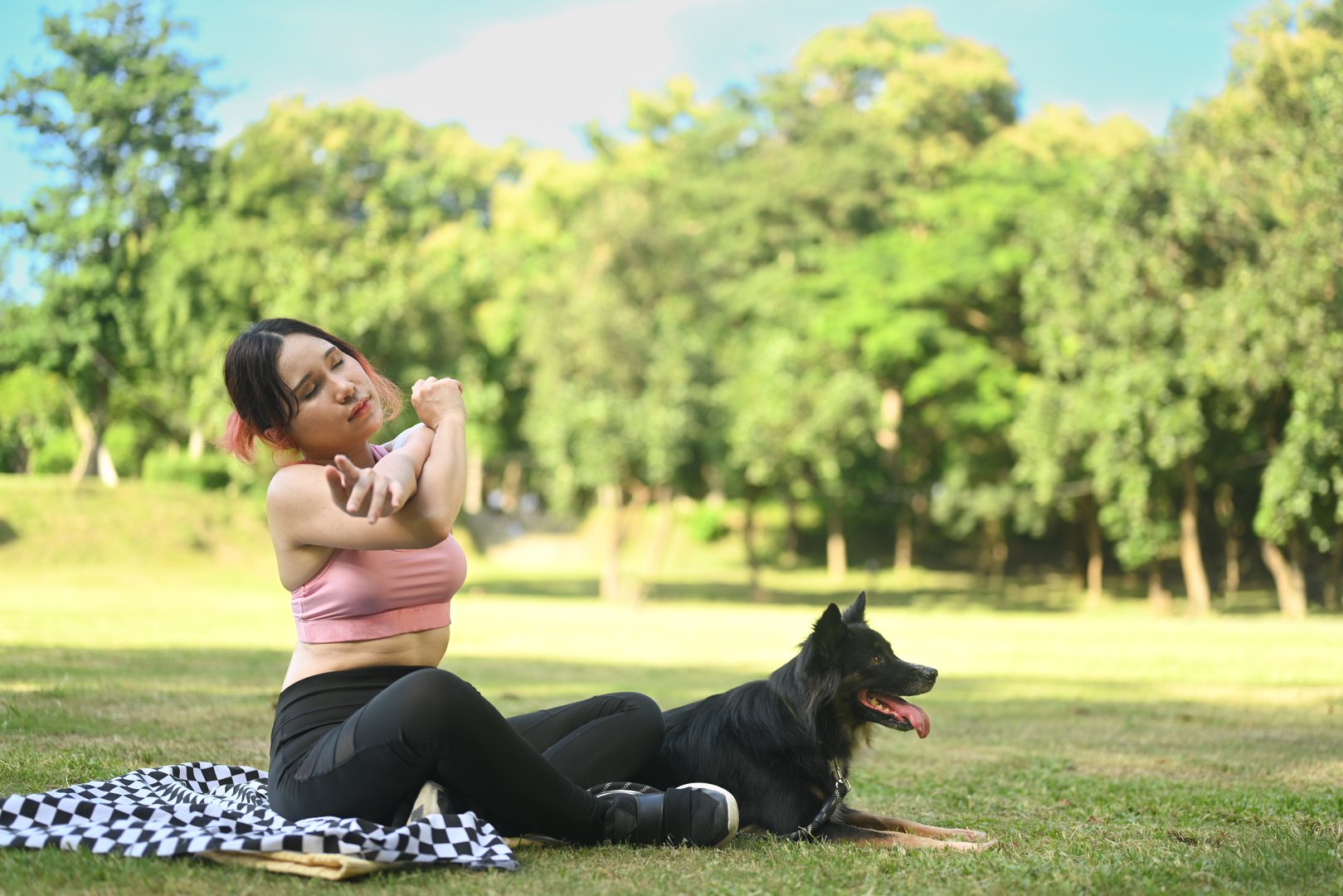 Young woman sitting on a blanket in a park with her border collie dog and stretching her arms after workout.