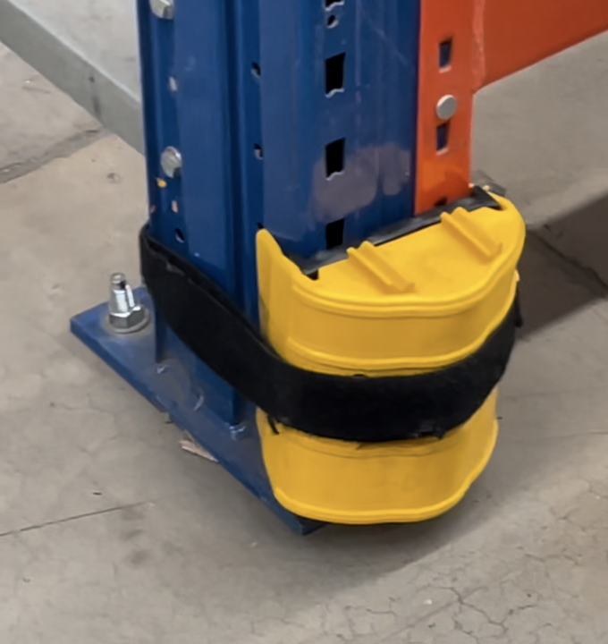 Warehouse racking with a yellow protective guard on the base of a blue upright, secured by a black strap.