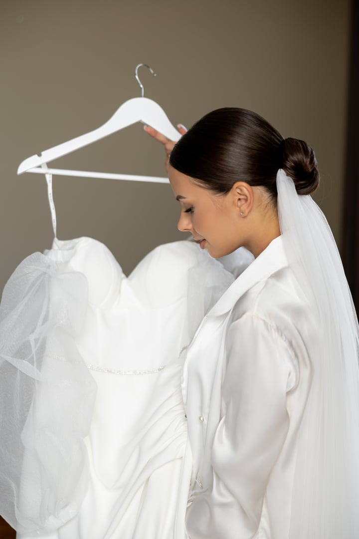 Beautiful bride wearing bathrobe and veil holding her white wedding dress on a hanger, smiling