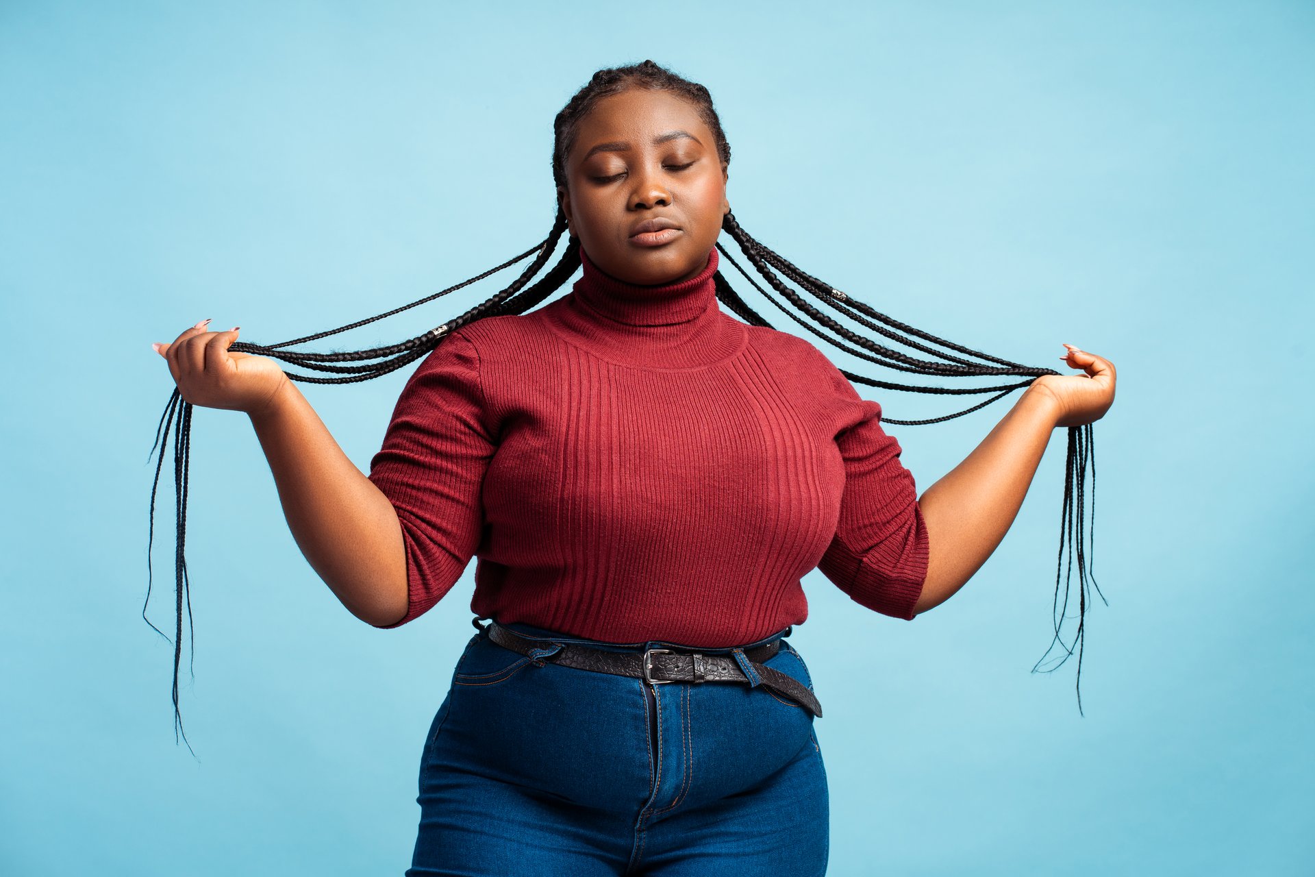 Cheerful beautiful African American woman with stylish braids, wearing casual clothes, red sweater and denim with closed eyes posing isolated on blue background. Hair care concept