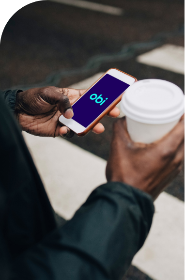 mask 4 | Iterators A person standing outside, holding a smartphone showing the "obi" logo on the screen in one hand and a white disposable coffee cup in the other.