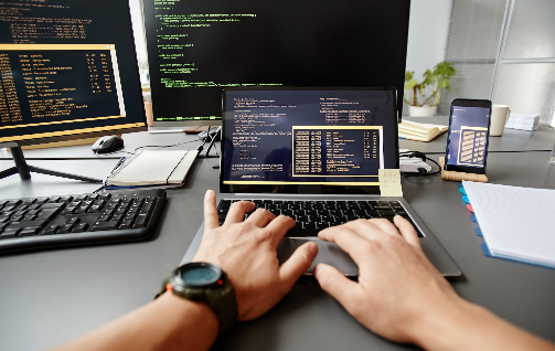 background-image-diverse-group-people-smiling-in-m-2025-03-05-05-12-39-utc-2 | Iterators Person working at a desk with multiple monitors displaying code, typing on a laptop in a modern office environment.