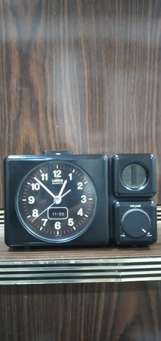 ***********
ساعة منبه مكتبية تناظرية (Annuity/Desk Alarm Clock) كلاسيكية سوداء اللون، مصممة بهيكل بلاستيكي مدمج ومقسمة إلى جزأين رئيسيين:
​الجانب الأيسر (جسم الساعة الرئيسي): يحتوي على قرص الساعة الدائري العقربي، وشاشة رقمية صغيرة مدمجة بالأسفل.
​الجانب الأيمن (وحدة الصوت): مخصص لنظام التنبيه الصوتي، ويحتوي على مكبر صوت (Speaker) في الأعلى ومفتاح دوار للتحكم في مستوى الصوت (Volume) في الأسفل.
​  قرص الساعة (The Dial)
​العلامة التجارية: يظهر في الجزء العلوي من القرص شعار LORUS QUARTZ، وهي علامة تجارية معروفة تابعة لشركة "سيكو" (Seiko) ومشهورة بدقة آلياتها المعتمدة على الكوارتز.
​الأرقام والعلامات: الأرقام من 1 إلى 12 مكتوبة بخط أبيض عريض وواضح على خلفية سوداء، وتوجد شرطات دقيقة حمراء/بيضاء تمثل الدقائق والثواني حول الإطار الداخلي.
​قراءة الوقت التناظري 
​عقرب التنبيه:  (وهو المخصص لضبط وقت الاستيقاظ).
​الشاشة الرقمية المدمجة: توجد نافذة رقمية (LCD) صغيرة في الجزء السفلي من القرص تعرض الوقت بصيغة رقمية وهي تشير بدقة إلى 11:55 (هناك فارق بسيط ومألوف في الساعات القديمة بين مطابقة العقارب الدقيقة والشاشة الرقمية المدمجة) وحدة التحكم والمميزات الخاصة
​مكبر الصوت (Speaker Grid): في الجزء العلوي الأيمن، محمي بشبكة دائرية معدنية لتوزيع صوت المنبه.
​مفتاح التحكم بالصوت (Volume Knob): في الجزء السفلي الأيمن، وهو مفتاح دوار ميكانيكي يبدأ من الحد الأدنى (min) وينتهي عند الحد الأقصى (max)، والمؤشر مضبوط حالياً بالقرب من الحد الأقصى (ما يعني أن صوت المنبه سيكون مرتفعاً جداً).
​زر الإيقاف/الإضاءة: يظهر في أعلى الساعة زر مستطيل ممتد، يُستخدم عادةً كزر غفوة (Snooze) أو لتشغيل إضاءة خلفية مؤقتة للقرص.
​الحالة العامة والخلفية
​الساعة  بحالة جيدة جداً ونظيفة، وتصميمها يعود غالباً إلى أواخر الثمانينيات أو فترة التسعينيات، وهو ما يُعرف بالتصاميم "الرديترية/الكلاسيكية" (Retro/Vintage).
