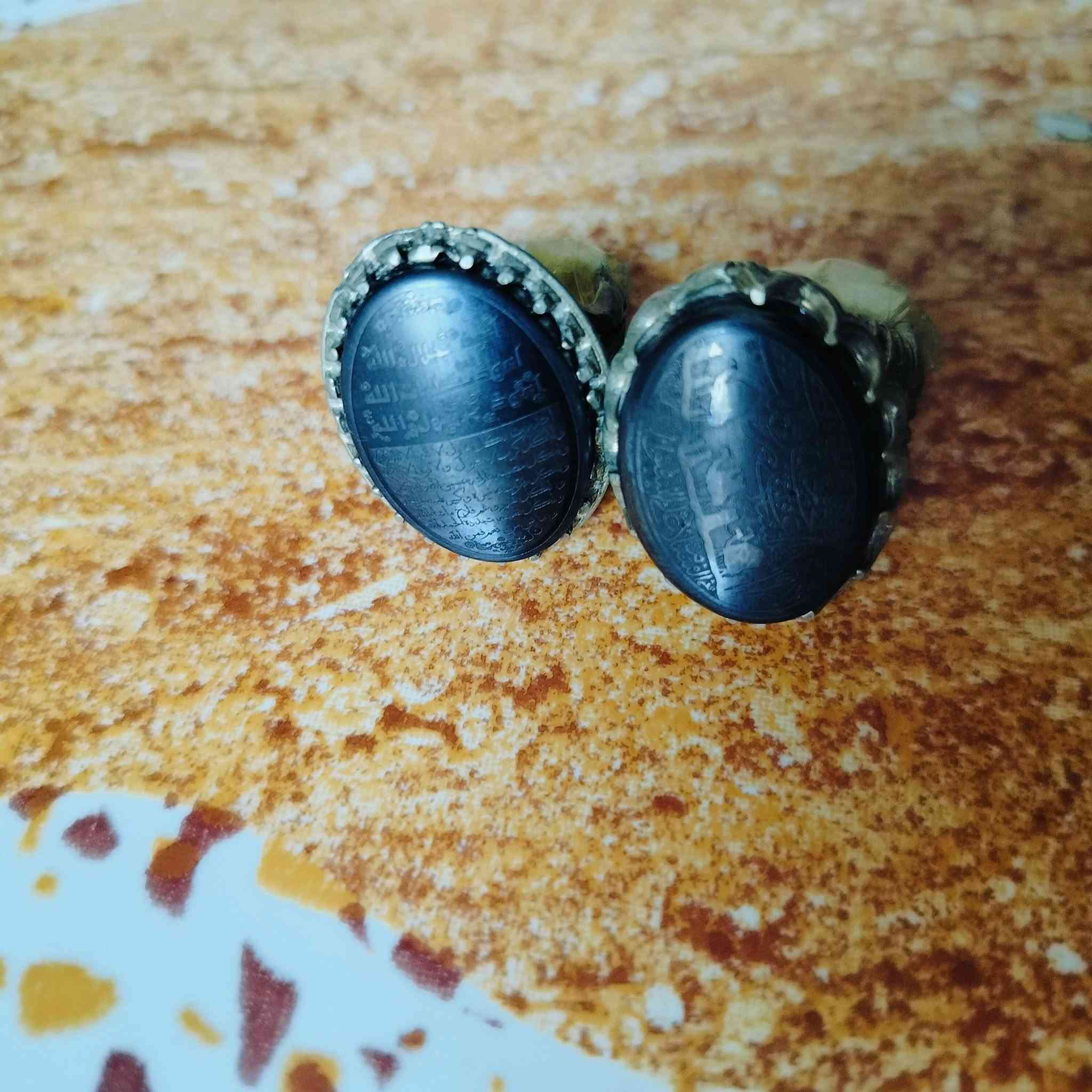 هذه المحابس تحتوي على أحجار من نوع الحديد الصيني (Hematite)، وهو نوع شائع ومشهور بنقش النقوش والأدعية والآيات الشريفة:
الخاتم على اليسار:
​منقوش عليه حرز الجلالات السبع، وهو نقش معروف ومأثور يُكتب على فص الحديد الصيني، ويتكون من السطور التالية:
​السطر الأول: أعوذ بجلال الله.
​السطر الثاني: أعوذ بكلمات الله.
​السطر الثالث: أعوذ برسول الله.
​باقي الأسطر: عبارات التوسل والتوكل المتممة للحرز مثل:
​"آمنت بالله وكتبه ورسله"
​"إني واثق بالله ورسله"
​"أشهد أن لا إله إلا الله مخلصاً"
​الخاتم على اليمين:
​يحتوي على نقش دعاء وتوسل بأهل البيت (عليهم السلام) مع أشكال رمزية (مثل كف أبي الفضل العباس أو السيف):
​عبارة "يا علي" أو "يا أبا الفضل العباس" في الجانب.
​أدعية للتحصين والسلامة وطلب الحاجة.
هذه المحابس تحتوي على أحجار من نوع الحديد الصيني (Hematite)، وهو نوع شائع ومشهور بنقش النقوش والأدعية والآيات الشريفة: والسعر خاص.. على هذا الرقم..  
***********
