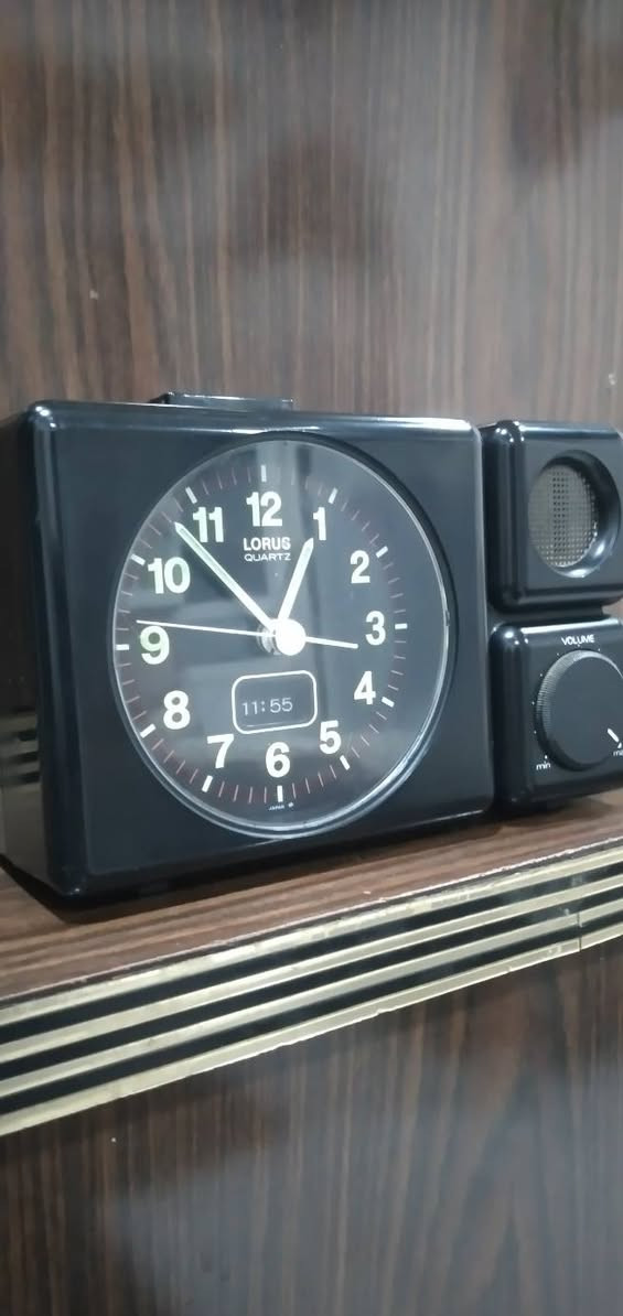 ***********
ساعة منبه مكتبية تناظرية (Annuity/Desk Alarm Clock) كلاسيكية سوداء اللون، مصممة بهيكل بلاستيكي مدمج ومقسمة إلى جزأين رئيسيين:
​الجانب الأيسر (جسم الساعة الرئيسي): يحتوي على قرص الساعة الدائري العقربي، وشاشة رقمية صغيرة مدمجة بالأسفل.
​الجانب الأيمن (وحدة الصوت): مخصص لنظام التنبيه الصوتي، ويحتوي على مكبر صوت (Speaker) في الأعلى ومفتاح دوار للتحكم في مستوى الصوت (Volume) في الأسفل.
​  قرص الساعة (The Dial)
​العلامة التجارية: يظهر في الجزء العلوي من القرص شعار LORUS QUARTZ، وهي علامة تجارية معروفة تابعة لشركة "سيكو" (Seiko) ومشهورة بدقة آلياتها المعتمدة على الكوارتز.
​الأرقام والعلامات: الأرقام من 1 إلى 12 مكتوبة بخط أبيض عريض وواضح على خلفية سوداء، وتوجد شرطات دقيقة حمراء/بيضاء تمثل الدقائق والثواني حول الإطار الداخلي.
​قراءة الوقت التناظري 
​عقرب التنبيه:  (وهو المخصص لضبط وقت الاستيقاظ).
​الشاشة الرقمية المدمجة: توجد نافذة رقمية (LCD) صغيرة في الجزء السفلي من القرص تعرض الوقت بصيغة رقمية وهي تشير بدقة إلى 11:55 (هناك فارق بسيط ومألوف في الساعات القديمة بين مطابقة العقارب الدقيقة والشاشة الرقمية المدمجة) وحدة التحكم والمميزات الخاصة
​مكبر الصوت (Speaker Grid): في الجزء العلوي الأيمن، محمي بشبكة دائرية معدنية لتوزيع صوت المنبه.
​مفتاح التحكم بالصوت (Volume Knob): في الجزء السفلي الأيمن، وهو مفتاح دوار ميكانيكي يبدأ من الحد الأدنى (min) وينتهي عند الحد الأقصى (max)، والمؤشر مضبوط حالياً بالقرب من الحد الأقصى (ما يعني أن صوت المنبه سيكون مرتفعاً جداً).
​زر الإيقاف/الإضاءة: يظهر في أعلى الساعة زر مستطيل ممتد، يُستخدم عادةً كزر غفوة (Snooze) أو لتشغيل إضاءة خلفية مؤقتة للقرص.
​الحالة العامة والخلفية
​الساعة  بحالة جيدة جداً ونظيفة، وتصميمها يعود غالباً إلى أواخر الثمانينيات أو فترة التسعينيات، وهو ما يُعرف بالتصاميم "الرديترية/الكلاسيكية" (Retro/Vintage).
