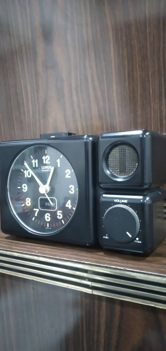***********
ساعة منبه مكتبية تناظرية (Annuity/Desk Alarm Clock) كلاسيكية سوداء اللون، مصممة بهيكل بلاستيكي مدمج ومقسمة إلى جزأين رئيسيين:
​الجانب الأيسر (جسم الساعة الرئيسي): يحتوي على قرص الساعة الدائري العقربي، وشاشة رقمية صغيرة مدمجة بالأسفل.
​الجانب الأيمن (وحدة الصوت): مخصص لنظام التنبيه الصوتي، ويحتوي على مكبر صوت (Speaker) في الأعلى ومفتاح دوار للتحكم في مستوى الصوت (Volume) في الأسفل.
​  قرص الساعة (The Dial)
​العلامة التجارية: يظهر في الجزء العلوي من القرص شعار LORUS QUARTZ، وهي علامة تجارية معروفة تابعة لشركة "سيكو" (Seiko) ومشهورة بدقة آلياتها المعتمدة على الكوارتز.
​الأرقام والعلامات: الأرقام من 1 إلى 12 مكتوبة بخط أبيض عريض وواضح على خلفية سوداء، وتوجد شرطات دقيقة حمراء/بيضاء تمثل الدقائق والثواني حول الإطار الداخلي.
​قراءة الوقت التناظري 
​عقرب التنبيه:  (وهو المخصص لضبط وقت الاستيقاظ).
​الشاشة الرقمية المدمجة: توجد نافذة رقمية (LCD) صغيرة في الجزء السفلي من القرص تعرض الوقت بصيغة رقمية وهي تشير بدقة إلى 11:55 (هناك فارق بسيط ومألوف في الساعات القديمة بين مطابقة العقارب الدقيقة والشاشة الرقمية المدمجة) وحدة التحكم والمميزات الخاصة
​مكبر الصوت (Speaker Grid): في الجزء العلوي الأيمن، محمي بشبكة دائرية معدنية لتوزيع صوت المنبه.
​مفتاح التحكم بالصوت (Volume Knob): في الجزء السفلي الأيمن، وهو مفتاح دوار ميكانيكي يبدأ من الحد الأدنى (min) وينتهي عند الحد الأقصى (max)، والمؤشر مضبوط حالياً بالقرب من الحد الأقصى (ما يعني أن صوت المنبه سيكون مرتفعاً جداً).
​زر الإيقاف/الإضاءة: يظهر في أعلى الساعة زر مستطيل ممتد، يُستخدم عادةً كزر غفوة (Snooze) أو لتشغيل إضاءة خلفية مؤقتة للقرص.
​الحالة العامة والخلفية
​الساعة  بحالة جيدة جداً ونظيفة، وتصميمها يعود غالباً إلى أواخر الثمانينيات أو فترة التسعينيات، وهو ما يُعرف بالتصاميم "الرديترية/الكلاسيكية" (Retro/Vintage).
