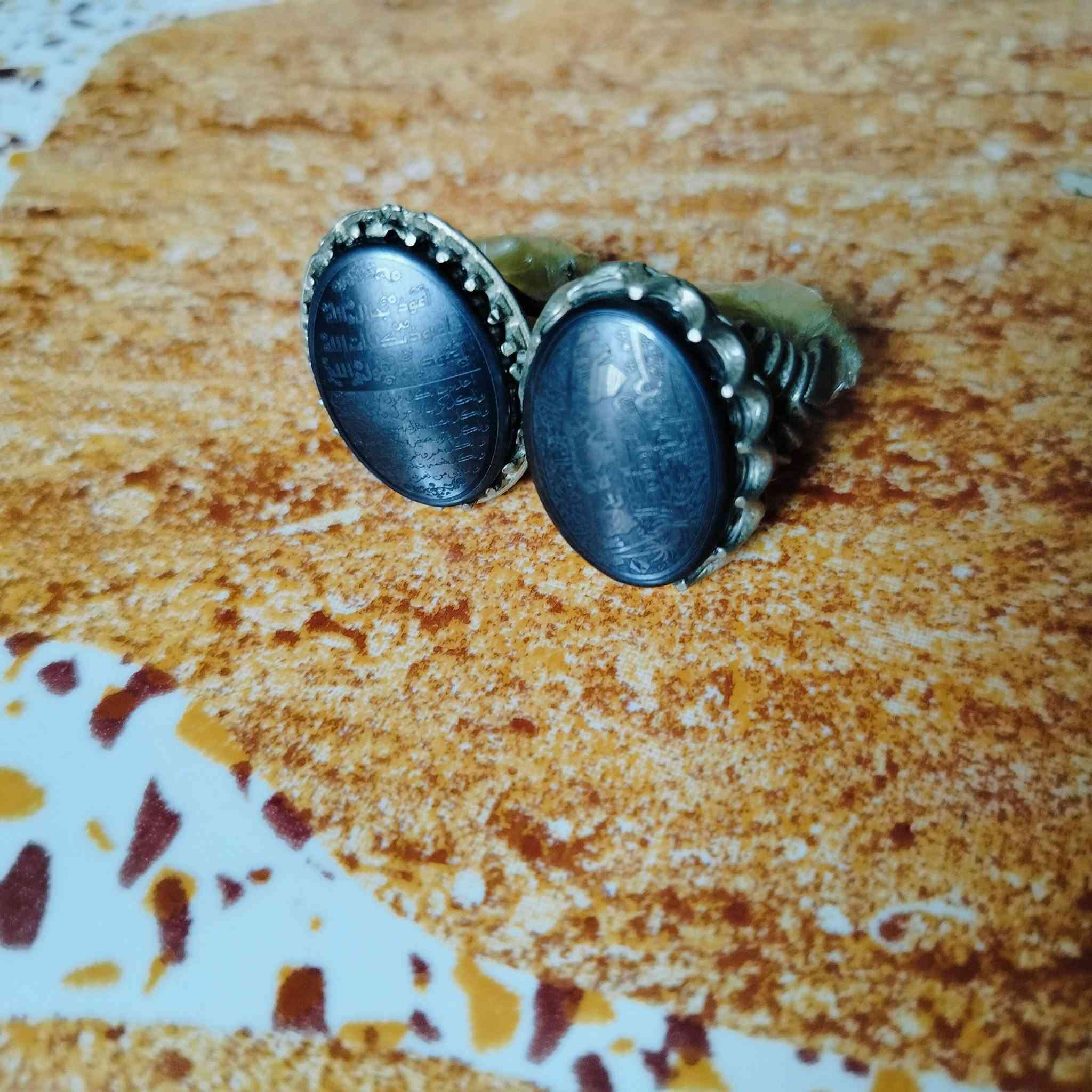 هذه المحابس تحتوي على أحجار من نوع الحديد الصيني (Hematite)، وهو نوع شائع ومشهور بنقش النقوش والأدعية والآيات الشريفة:
الخاتم على اليسار:
​منقوش عليه حرز الجلالات السبع، وهو نقش معروف ومأثور يُكتب على فص الحديد الصيني، ويتكون من السطور التالية:
​السطر الأول: أعوذ بجلال الله.
​السطر الثاني: أعوذ بكلمات الله.
​السطر الثالث: أعوذ برسول الله.
​باقي الأسطر: عبارات التوسل والتوكل المتممة للحرز مثل:
​"آمنت بالله وكتبه ورسله"
​"إني واثق بالله ورسله"
​"أشهد أن لا إله إلا الله مخلصاً"
​الخاتم على اليمين:
​يحتوي على نقش دعاء وتوسل بأهل البيت (عليهم السلام) مع أشكال رمزية (مثل كف أبي الفضل العباس أو السيف):
​عبارة "يا علي" أو "يا أبا الفضل العباس" في الجانب.
​أدعية للتحصين والسلامة وطلب الحاجة.
هذه المحابس تحتوي على أحجار من نوع الحديد الصيني (Hematite)، وهو نوع شائع ومشهور بنقش النقوش والأدعية والآيات الشريفة: والسعر خاص.. على هذا الرقم..  
***********
