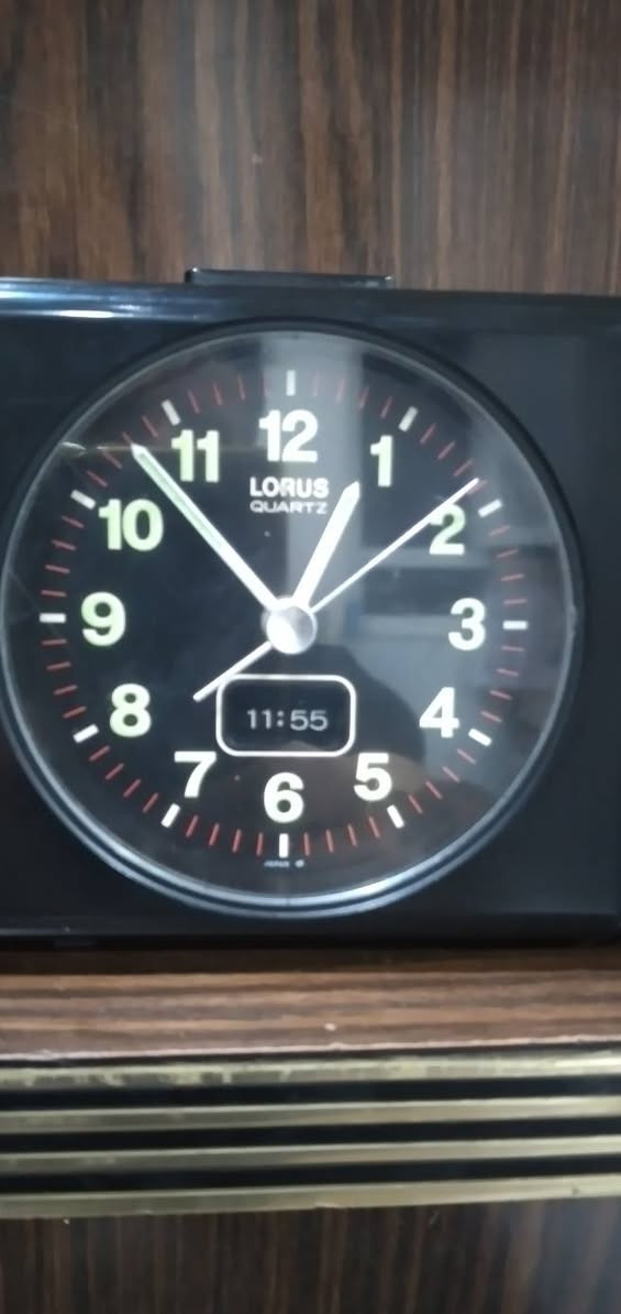 ***********
ساعة منبه مكتبية تناظرية (Annuity/Desk Alarm Clock) كلاسيكية سوداء اللون، مصممة بهيكل بلاستيكي مدمج ومقسمة إلى جزأين رئيسيين:
​الجانب الأيسر (جسم الساعة الرئيسي): يحتوي على قرص الساعة الدائري العقربي، وشاشة رقمية صغيرة مدمجة بالأسفل.
​الجانب الأيمن (وحدة الصوت): مخصص لنظام التنبيه الصوتي، ويحتوي على مكبر صوت (Speaker) في الأعلى ومفتاح دوار للتحكم في مستوى الصوت (Volume) في الأسفل.
​  قرص الساعة (The Dial)
​العلامة التجارية: يظهر في الجزء العلوي من القرص شعار LORUS QUARTZ، وهي علامة تجارية معروفة تابعة لشركة "سيكو" (Seiko) ومشهورة بدقة آلياتها المعتمدة على الكوارتز.
​الأرقام والعلامات: الأرقام من 1 إلى 12 مكتوبة بخط أبيض عريض وواضح على خلفية سوداء، وتوجد شرطات دقيقة حمراء/بيضاء تمثل الدقائق والثواني حول الإطار الداخلي.
​قراءة الوقت التناظري 
​عقرب التنبيه:  (وهو المخصص لضبط وقت الاستيقاظ).
​الشاشة الرقمية المدمجة: توجد نافذة رقمية (LCD) صغيرة في الجزء السفلي من القرص تعرض الوقت بصيغة رقمية وهي تشير بدقة إلى 11:55 (هناك فارق بسيط ومألوف في الساعات القديمة بين مطابقة العقارب الدقيقة والشاشة الرقمية المدمجة) وحدة التحكم والمميزات الخاصة
​مكبر الصوت (Speaker Grid): في الجزء العلوي الأيمن، محمي بشبكة دائرية معدنية لتوزيع صوت المنبه.
​مفتاح التحكم بالصوت (Volume Knob): في الجزء السفلي الأيمن، وهو مفتاح دوار ميكانيكي يبدأ من الحد الأدنى (min) وينتهي عند الحد الأقصى (max)، والمؤشر مضبوط حالياً بالقرب من الحد الأقصى (ما يعني أن صوت المنبه سيكون مرتفعاً جداً).
​زر الإيقاف/الإضاءة: يظهر في أعلى الساعة زر مستطيل ممتد، يُستخدم عادةً كزر غفوة (Snooze) أو لتشغيل إضاءة خلفية مؤقتة للقرص.
​الحالة العامة والخلفية
​الساعة  بحالة جيدة جداً ونظيفة، وتصميمها يعود غالباً إلى أواخر الثمانينيات أو فترة التسعينيات، وهو ما يُعرف بالتصاميم "الرديترية/الكلاسيكية" (Retro/Vintage).
