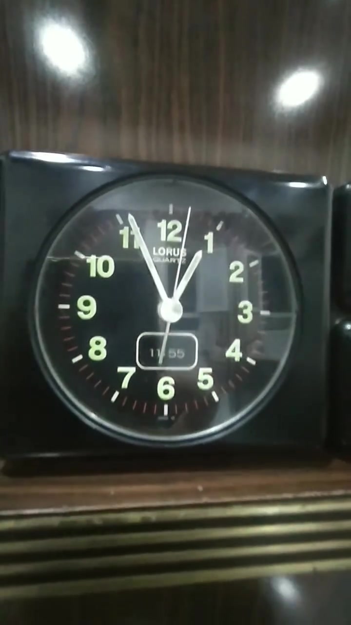 ***********
ساعة منبه مكتبية تناظرية (Annuity/Desk Alarm Clock) كلاسيكية سوداء اللون، مصممة بهيكل بلاستيكي مدمج ومقسمة إلى جزأين رئيسيين:
​الجانب الأيسر (جسم الساعة الرئيسي): يحتوي على قرص الساعة الدائري العقربي، وشاشة رقمية صغيرة مدمجة بالأسفل.
​الجانب الأيمن (وحدة الصوت): مخصص لنظام التنبيه الصوتي، ويحتوي على مكبر صوت (Speaker) في الأعلى ومفتاح دوار للتحكم في مستوى الصوت (Volume) في الأسفل.
​  قرص الساعة (The Dial)
​العلامة التجارية: يظهر في الجزء العلوي من القرص شعار LORUS QUARTZ، وهي علامة تجارية معروفة تابعة لشركة "سيكو" (Seiko) ومشهورة بدقة آلياتها المعتمدة على الكوارتز.
​الأرقام والعلامات: الأرقام من 1 إلى 12 مكتوبة بخط أبيض عريض وواضح على خلفية سوداء، وتوجد شرطات دقيقة حمراء/بيضاء تمثل الدقائق والثواني حول الإطار الداخلي.
​قراءة الوقت التناظري 
​عقرب التنبيه:  (وهو المخصص لضبط وقت الاستيقاظ).
​الشاشة الرقمية المدمجة: توجد نافذة رقمية (LCD) صغيرة في الجزء السفلي من القرص تعرض الوقت بصيغة رقمية وهي تشير بدقة إلى 11:55 (هناك فارق بسيط ومألوف في الساعات القديمة بين مطابقة العقارب الدقيقة والشاشة الرقمية المدمجة) وحدة التحكم والمميزات الخاصة
​مكبر الصوت (Speaker Grid): في الجزء العلوي الأيمن، محمي بشبكة دائرية معدنية لتوزيع صوت المنبه.
​مفتاح التحكم بالصوت (Volume Knob): في الجزء السفلي الأيمن، وهو مفتاح دوار ميكانيكي يبدأ من الحد الأدنى (min) وينتهي عند الحد الأقصى (max)، والمؤشر مضبوط حالياً بالقرب من الحد الأقصى (ما يعني أن صوت المنبه سيكون مرتفعاً جداً).
​زر الإيقاف/الإضاءة: يظهر في أعلى الساعة زر مستطيل ممتد، يُستخدم عادةً كزر غفوة (Snooze) أو لتشغيل إضاءة خلفية مؤقتة للقرص.
​الحالة العامة والخلفية
​الساعة  بحالة جيدة جداً ونظيفة، وتصميمها يعود غالباً إلى أواخر الثمانينيات أو فترة التسعينيات، وهو ما يُعرف بالتصاميم "الرديترية/الكلاسيكية" (Retro/Vintage).

