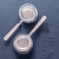 Two stainless steel European Hawthorne cocktail strainers on textured surface