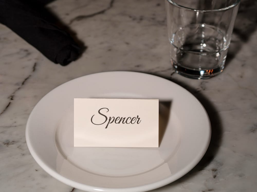 Place setting at a DougCo Social Dinner Club table — handwritten name card on a white plate, marble tabletop, glass tumbler