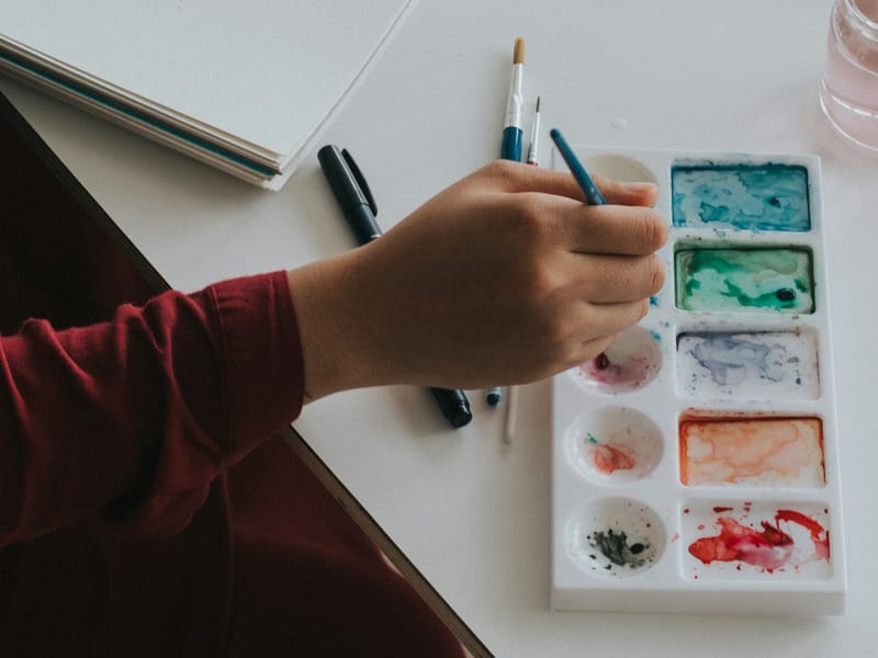 Illustrative image: a hand dipping a paintbrush into a watercolor palette