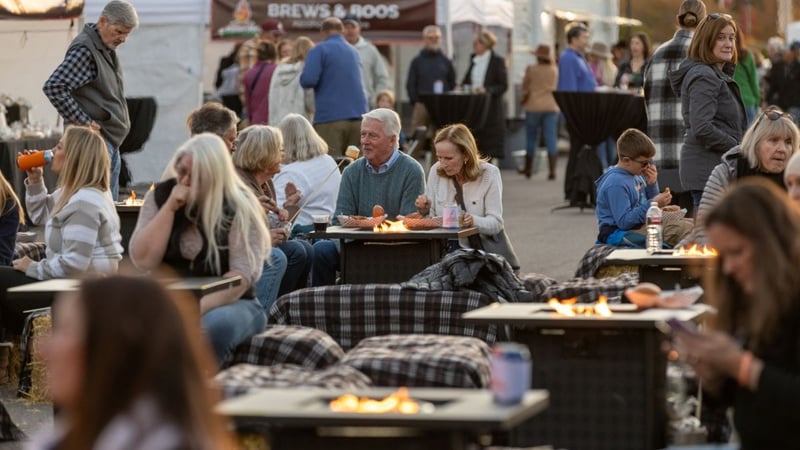 Crowd gathered around fire pit tables with a "Brews & Roos" banner at a past Bonfire BBQ & Brews