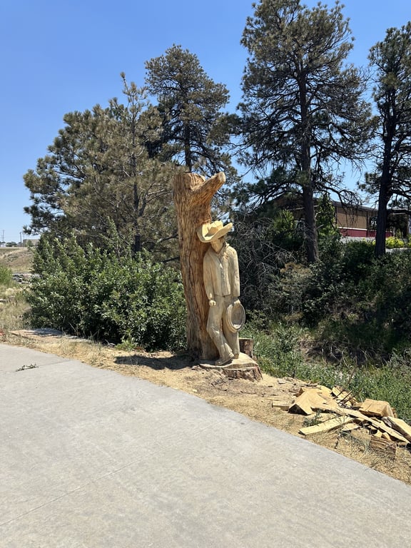 The Hangman's Gulch cowboy sculpture standing trailside in Castle Rock, with the paved trail in the foreground and pine trees behind.