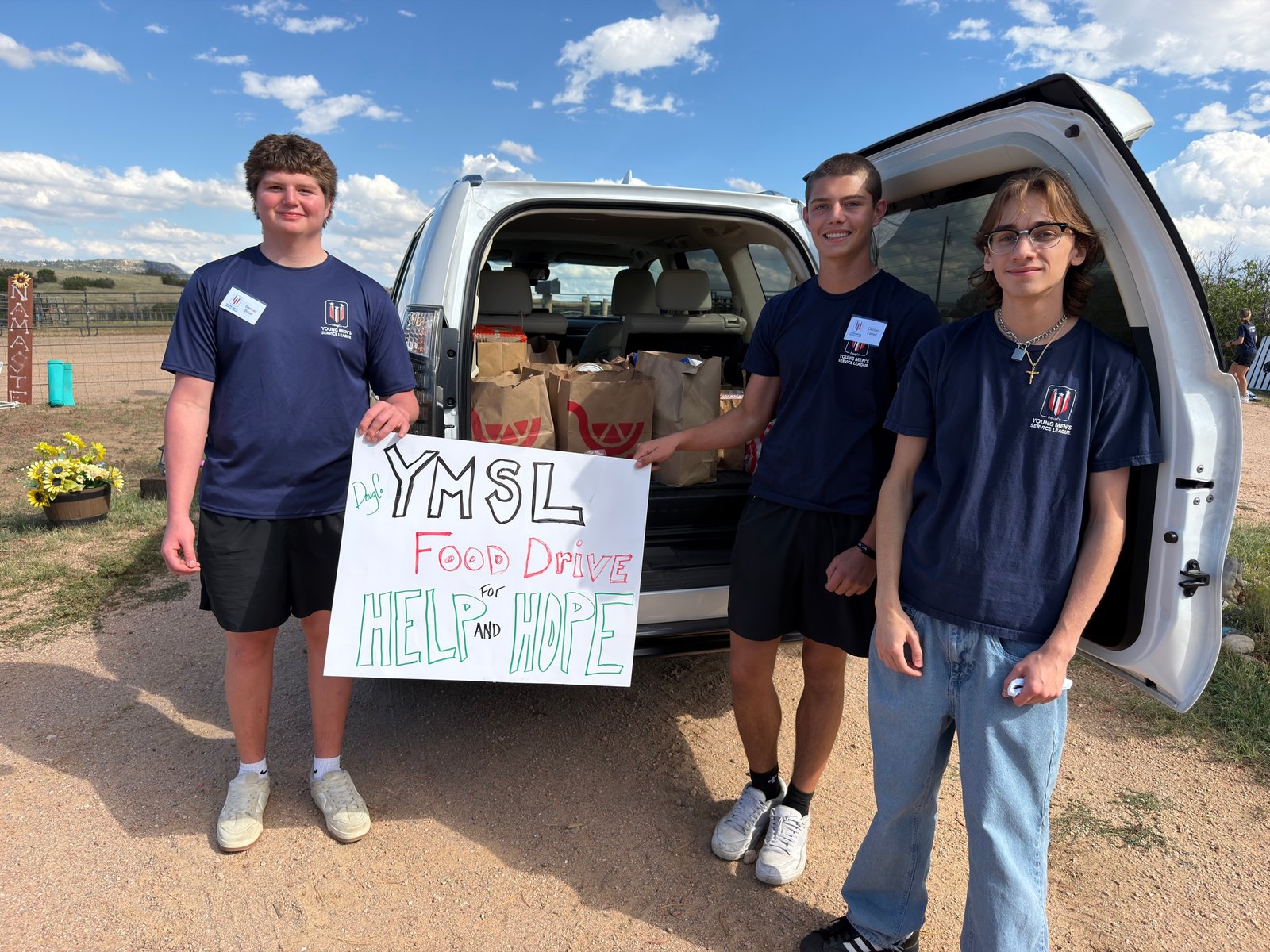 Three YMSL DougCo volunteers stand beside an SUV loaded with bagged food donations collected for Help & Hope Center