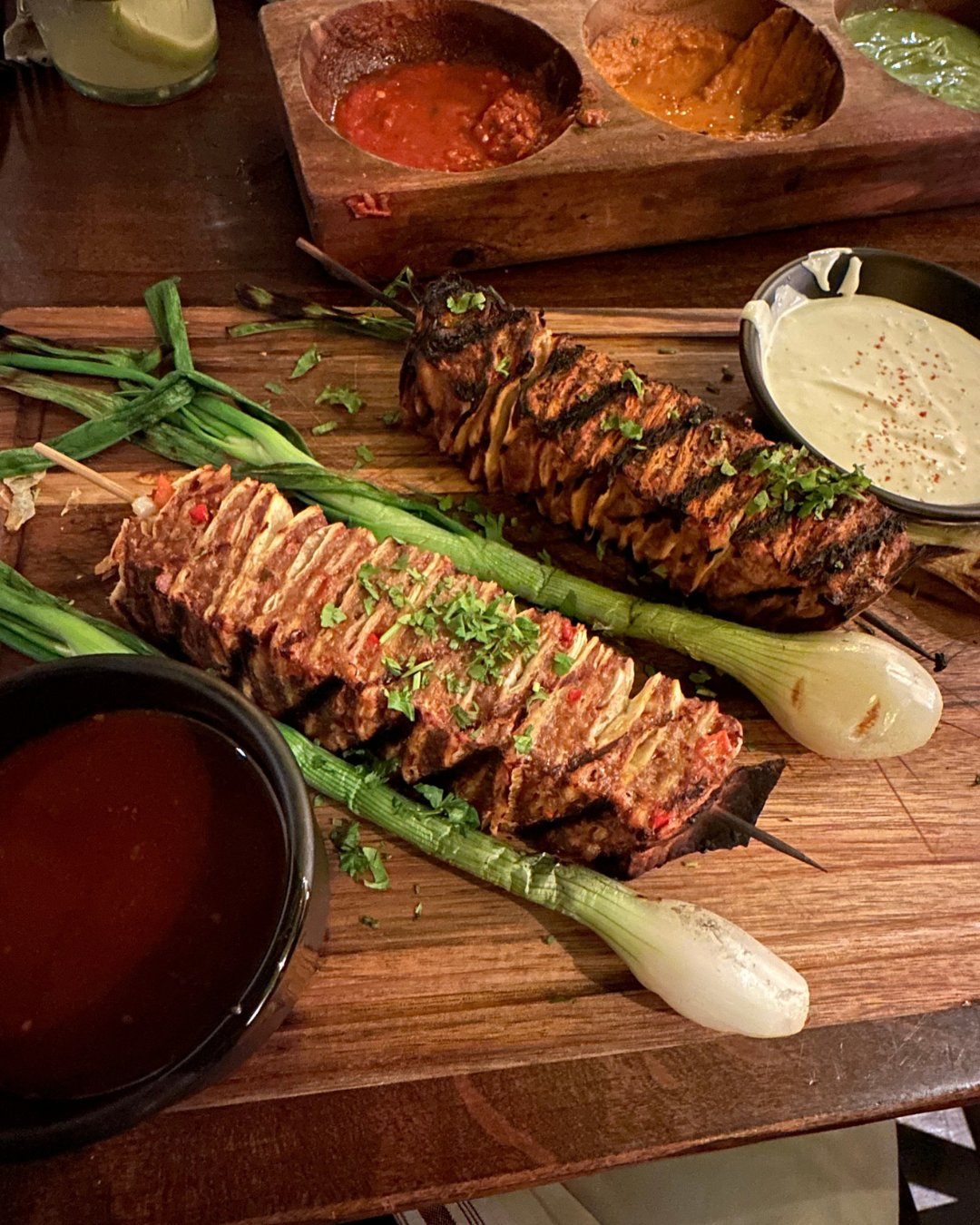Two hasselback-style birria dip skewers on a wood board with grilled green onions, consomme, a crema dip and a three-salsa flight, Bien Y Tu's Restaurant Week special