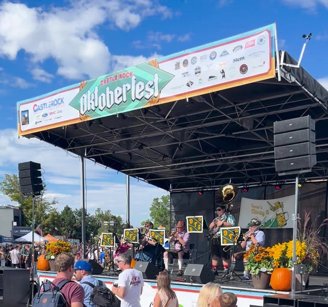 A polka band in lederhosen playing on the Castle Rock Oktoberfest stage under a banner, with pumpkins and sunflowers along the stage front and a crowd watching.