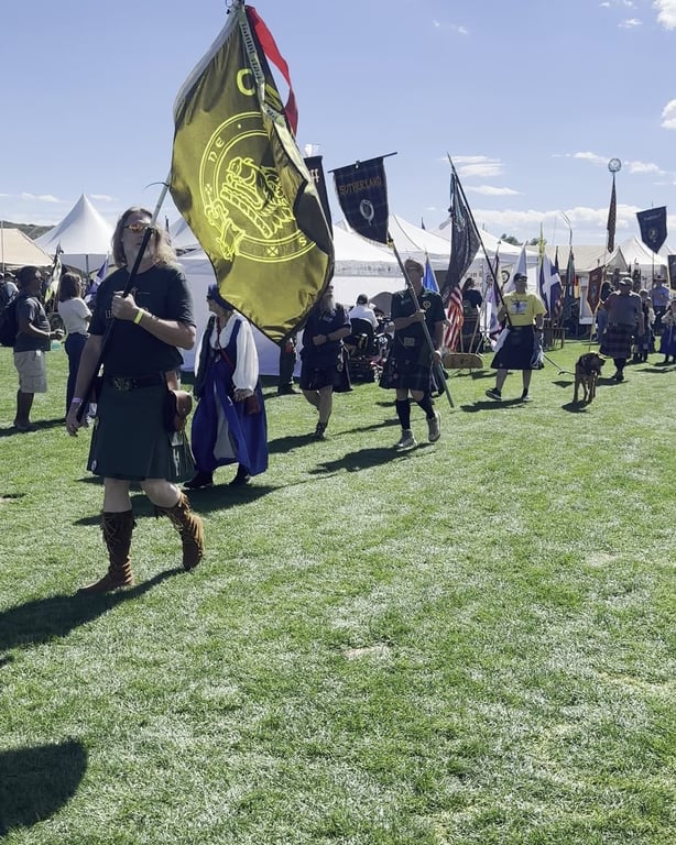 Clan members in kilts carrying banners across the field at the Colorado Highland Games