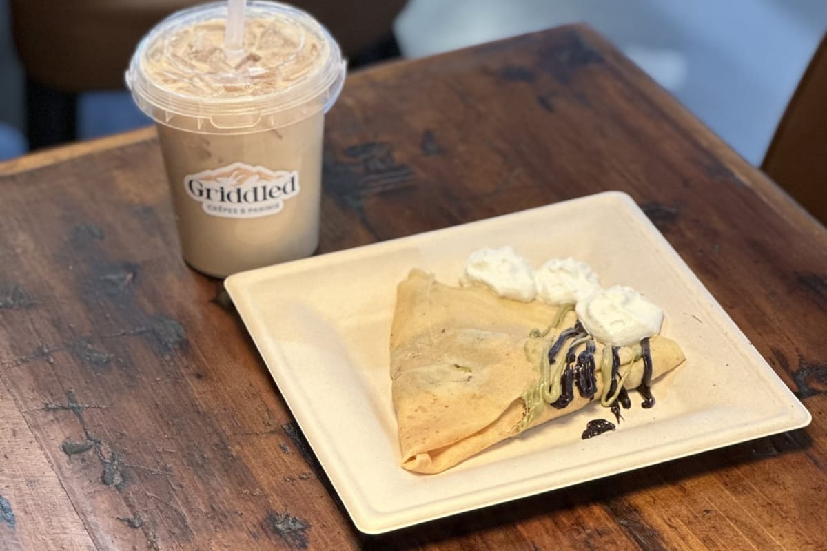 A chocolate-banana crepe with whipped cream and a branded Griddled iced latte on a wood table
