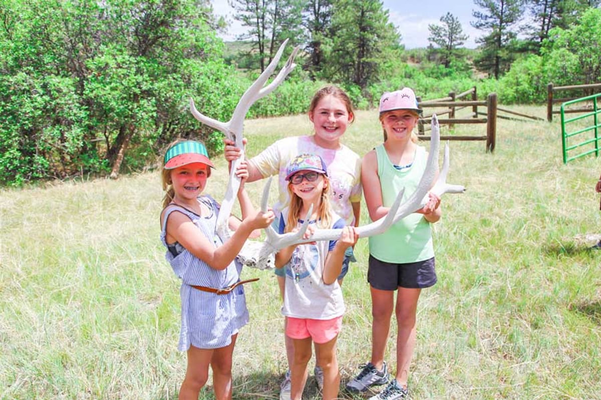 Four girls in a meadow at Camp Backcountry hold up shed elk antlers, with ponderosa pines and a wooden fence behind them