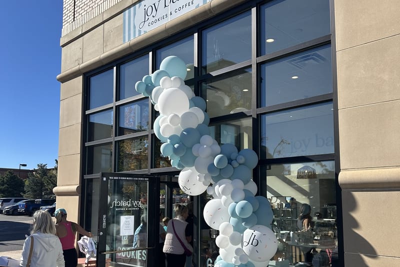Joy Batch Cookies & Coffee storefront in Highlands Ranch on opening day, Oct. 9, 2026, with a blue and white balloon arch and customers entering