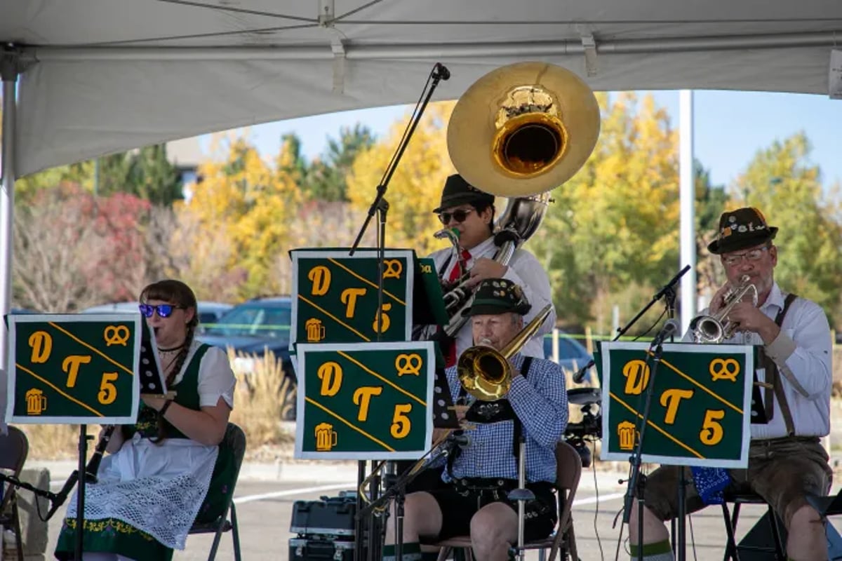 A four-piece brass band in lederhosen and dirndls plays under a tent at Lone Tree Oktoberfest with fall trees behind