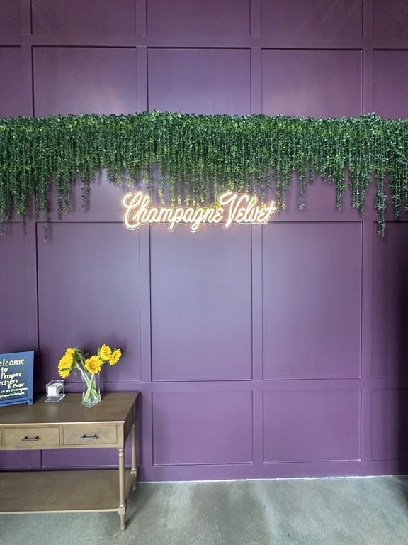 Champagne Velvet neon sign on plum-purple panel wall with hanging eucalyptus and welcome chalkboard