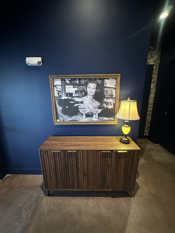Black-and-white portrait of woman pouring champagne in gold frame above wood console at Champagne Velvet Proper Kitchen & Bar