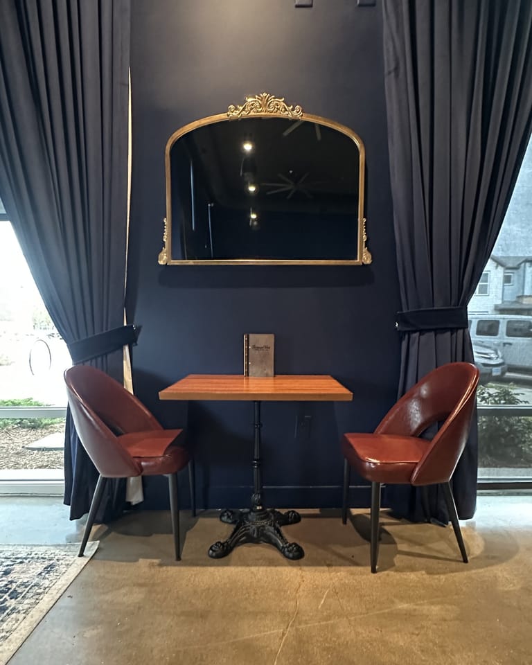Two-top by window with gold-framed mirror and burgundy chairs at Champagne Velvet Proper Kitchen & Bar