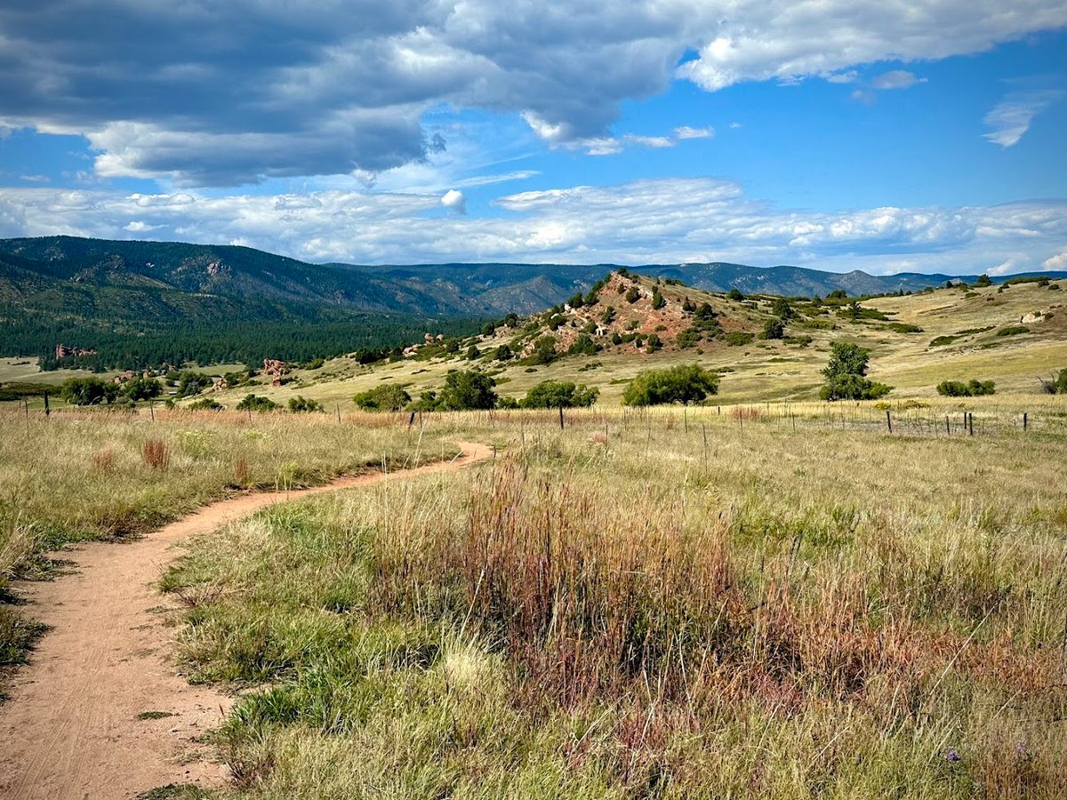 A dirt trail winding through tall grass at Sandstone Ranch Open Space in Larkspur, with red rock outcrops and the Front Range behind it.