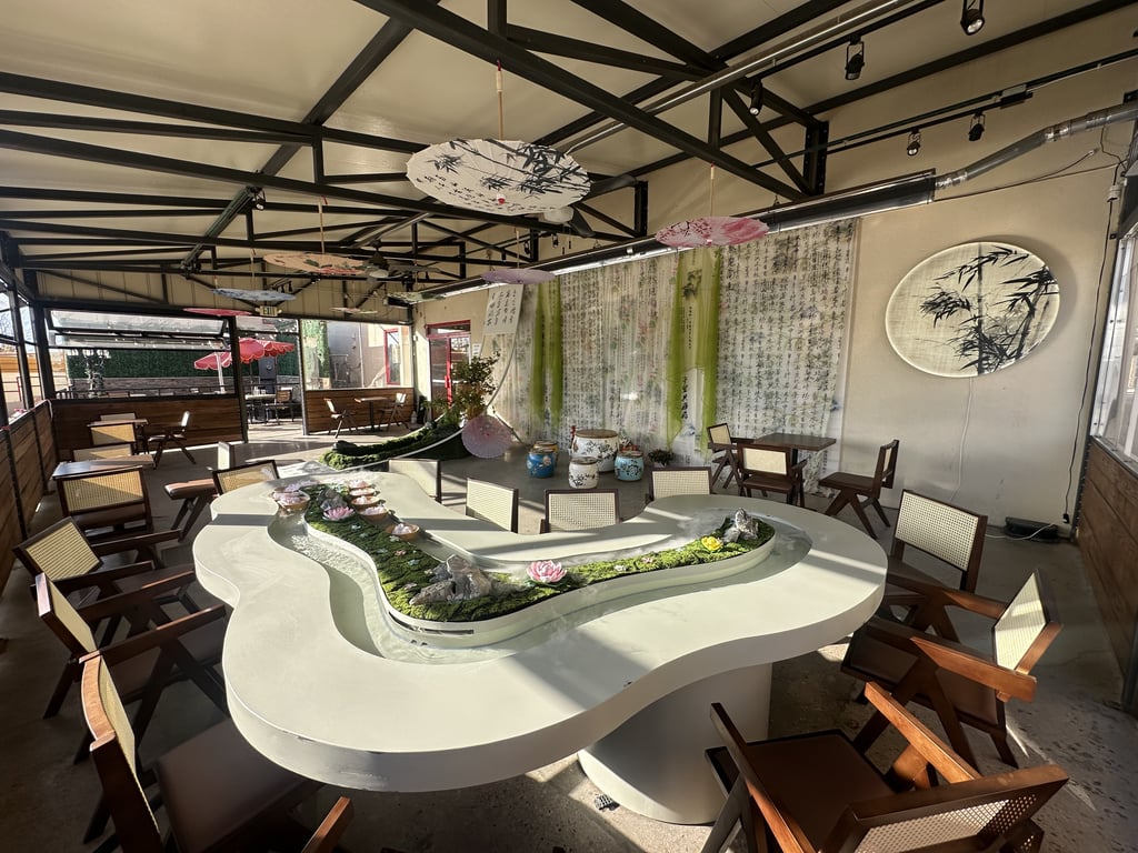 The white stream table with lotus bowls, pink and ink-painted paper parasols hanging overhead, and bamboo moon art on the wall of SereniTea's patio