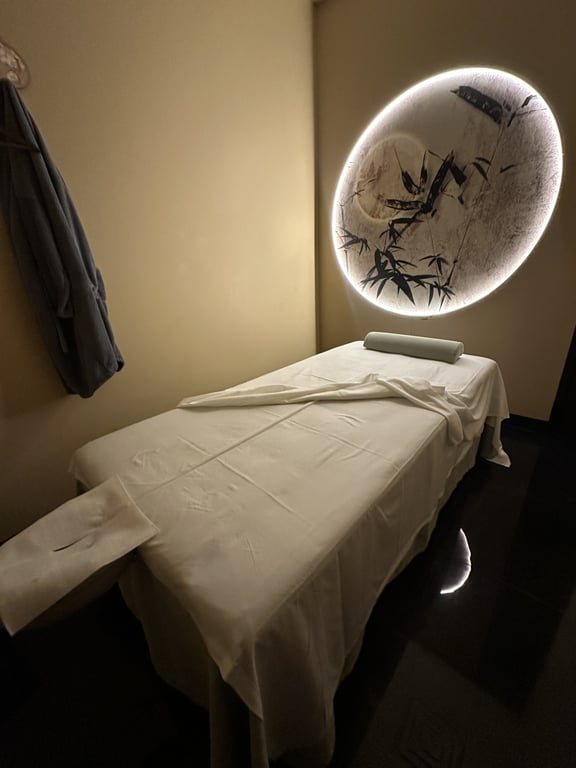 A treatment room at SereniTea Spa & Tea Lounge with a made-up massage table under a backlit round moon window painted with bamboo