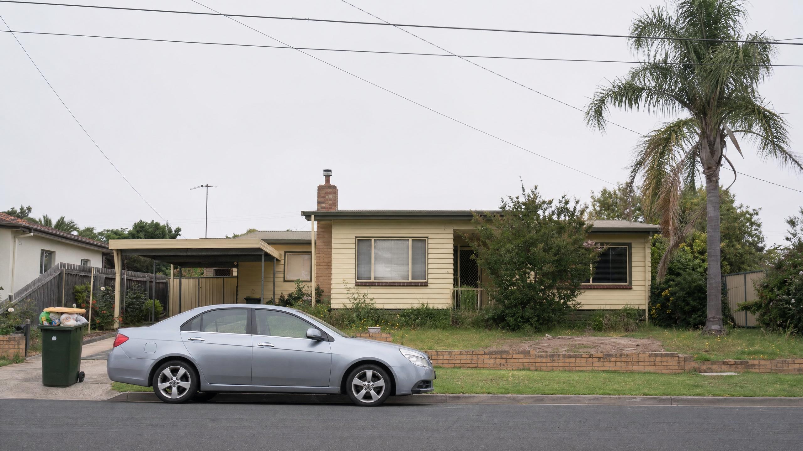 A yellow weatherboard house with a carport and palm tree under an overcast sky, a silver car parked out front