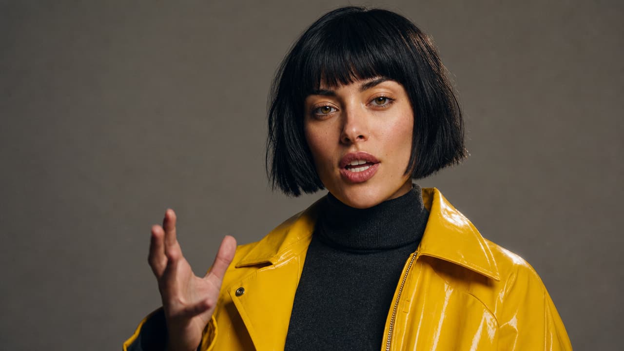 Woman with a jet-black bob in a chrome-yellow raincoat speaking mid-word to camera against a grey studio backdrop