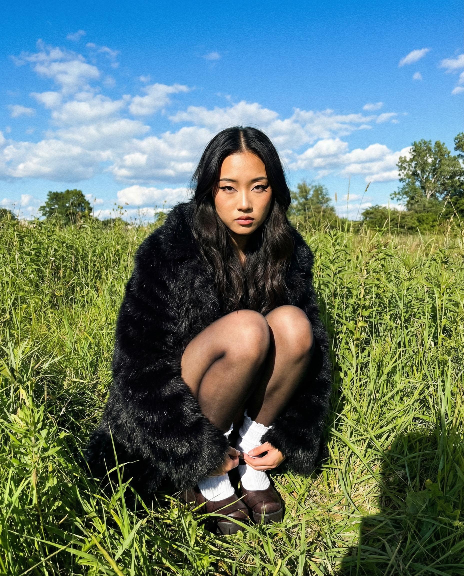 Woman in a black fur coat with winged eyeliner crouches in a grassy field under a blue sky with scattered clouds