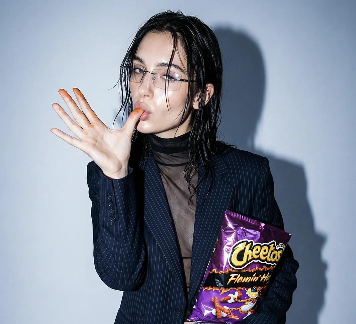 Woman in a pinstripe blazer licking her finger while holding a purple bag of Flamin' Hot Cheetos