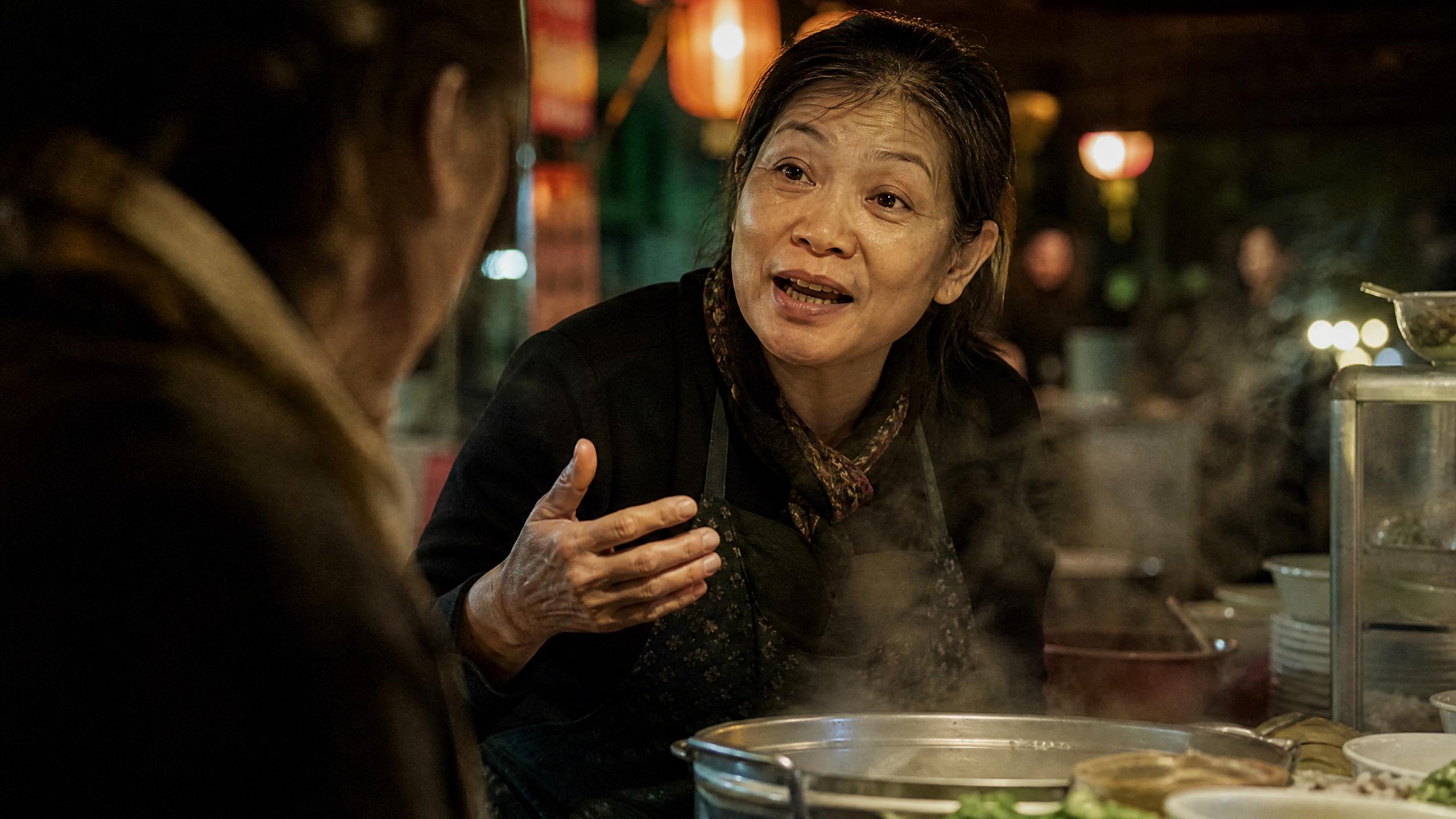 Vietnamese street-food cook mid-sentence at her lantern-lit night-market stall, hand raised, steam rising from a pot between her and a customer