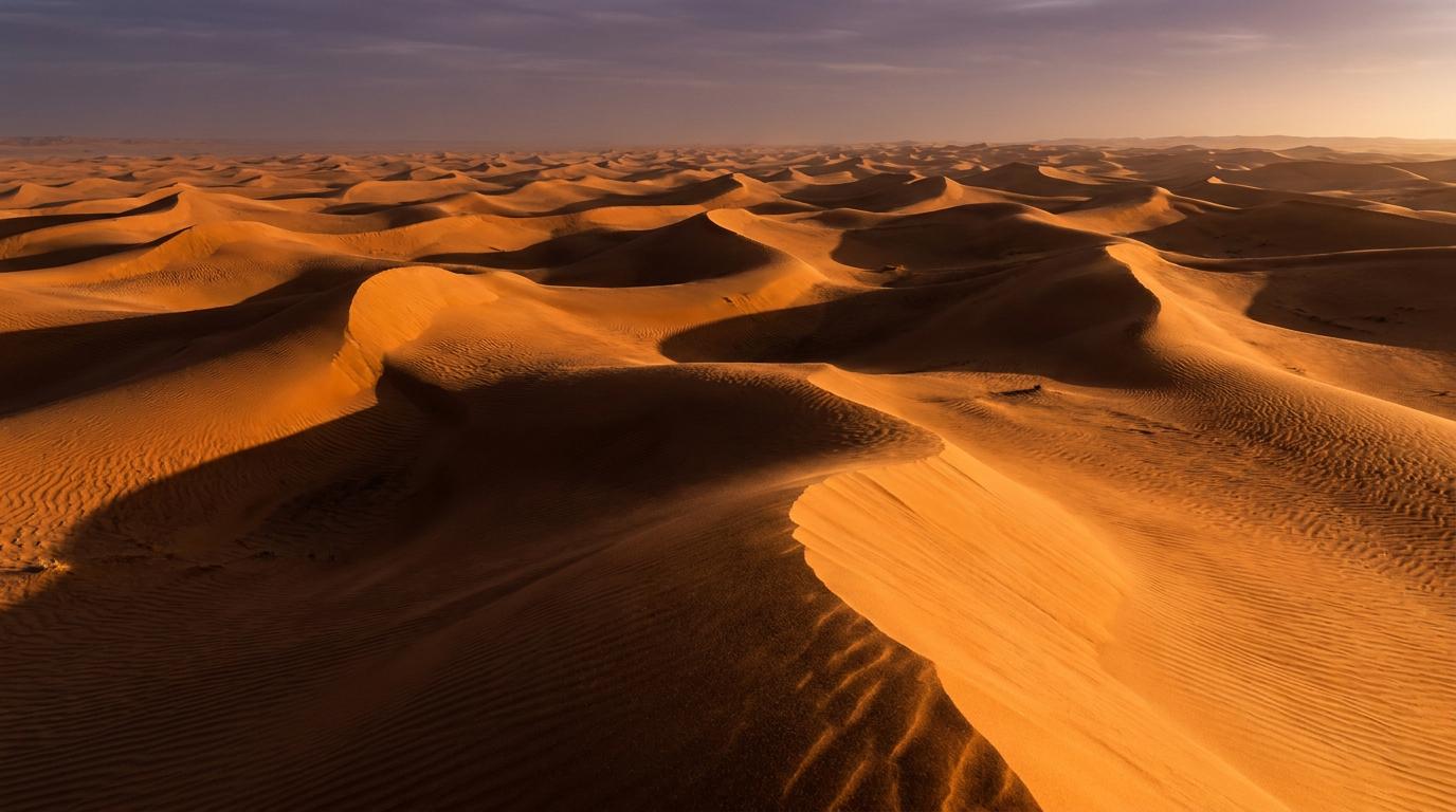 Rippled orange sand dunes stretching to the horizon under a hazy sky, with long shadows at sunset