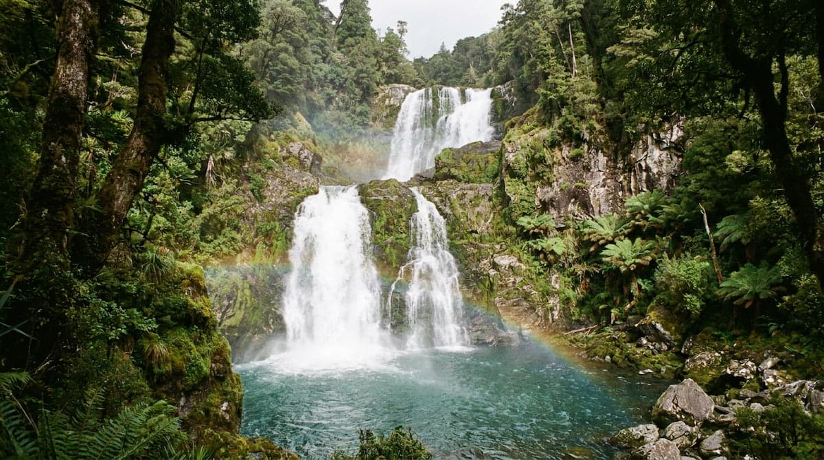 Multi-tiered waterfall cascading over mossy rocks into a blue-green pool, surrounded by lush forest with a faint rainbow