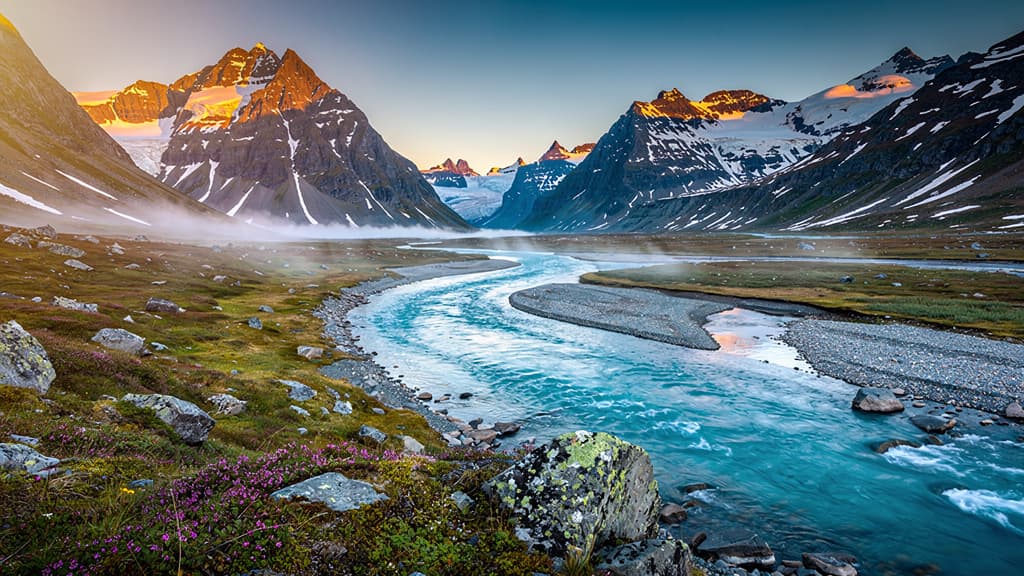A turquoise glacial river winds through Arctic tundra toward snow-dusted granite peaks at dawn.
