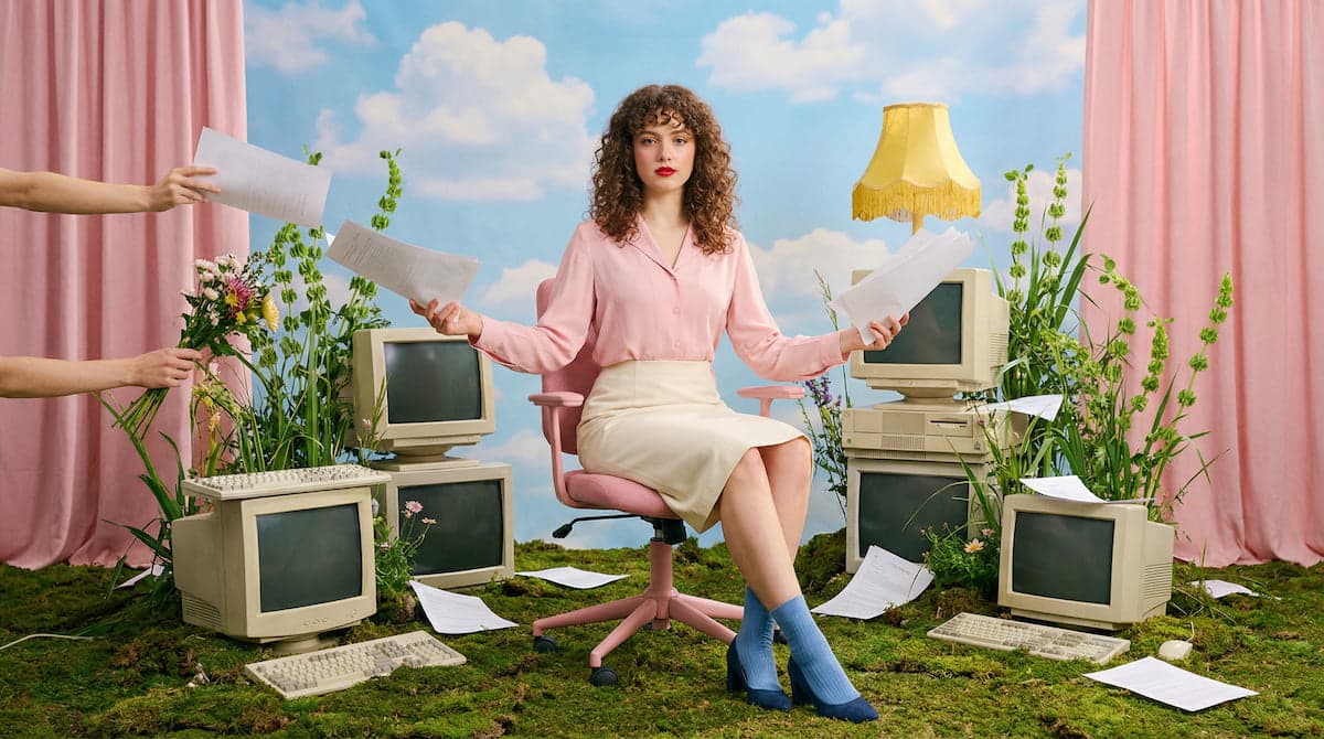 Woman in a pink blouse holding papers in a desk chair, surrounded by retro computers on a mossy floor